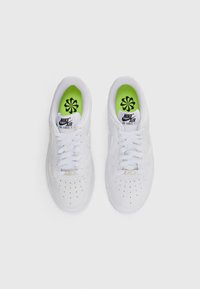 Un paio di sneakers Nike Air Force 1 bianche con punte perforate, lacci bianchi e soletta verde neon con loghi neri a forma di fiore.