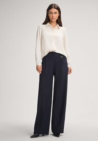 Vrouw in een crèmekleurige blouse met lange mouwen, ingestopt in een hoge taille met brede benen in marineblauwe broek en zwarte puntige schoenen.