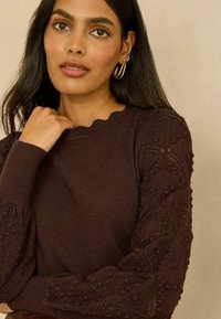 Pull en maille marron avec manches texturées à motif et encolure festonnée. Le modèle porte des boucles d'oreilles créoles dorées et a les cheveux longs et raides.