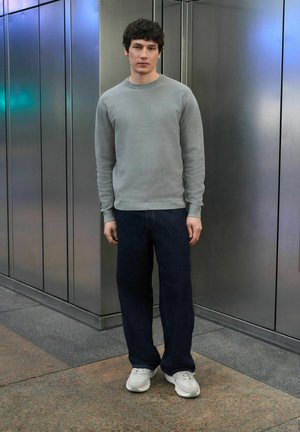Hellgrauer Pullover mit langen Ärmeln und strukturiertem Stoff, kombiniert mit dunkelblauen, locker sitzenden Jeans und hellgrauen Turnschuhen. Schlichtes urbanes Ambiente.