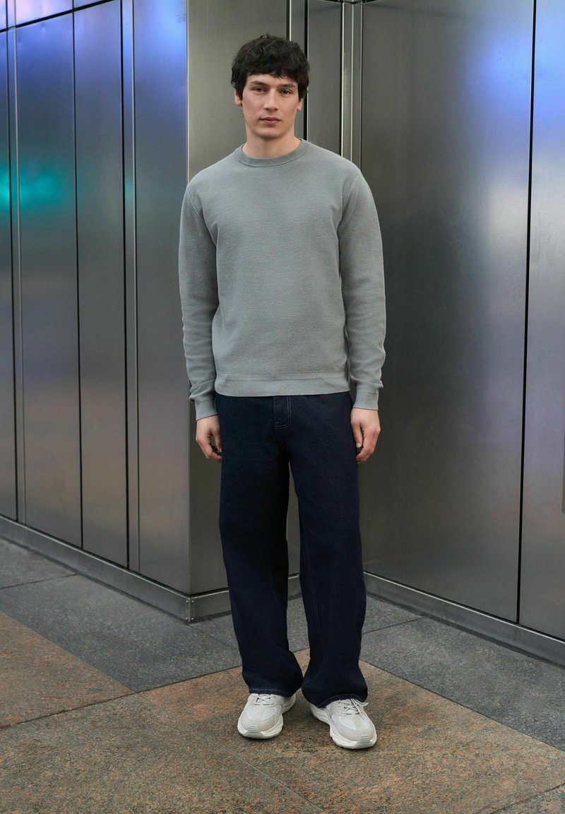 Sweat-shirt à manches longues gris clair avec tissu texturé, associé à un jean ample bleu foncé et des baskets gris clair. Cadre urbain simple.