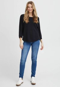 Schwarzer, strukturierter, locker sitzender Pullover mit Rundhalsausschnitt, kombiniert mit enganliegenden blauen Jeans und weißen Sneakers mit braunen Sohlen.