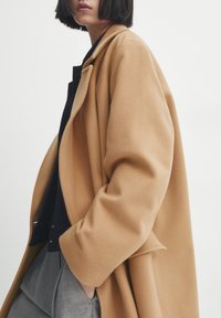 Manteau en laine couleur chameau, coupe ample et larges revers, porté sur un haut noir, accompagné d'un pantalon gris. Texture lisse.