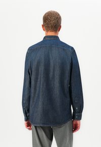 Man facing away wearing a dark blue denim long-sleeve shirt and light gray pants against a plain white background.
