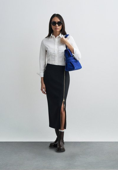 Μοντέλο με λευκό πουκάμισο με κουμπιά, μαύρη φούστα με φερμουάρ στο πλάι, μαύρα μποτάκια και κρατώντας μια μπλε τσάντα από σουέντ. Ορατές λεπτομέρειες υφής υφάσματος.