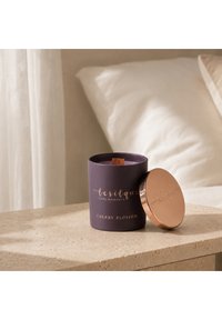 Lila Kirschblüten-Duftkerze mit Holzdocht und goldenem Deckel auf einem beigen Steinnachttisch neben weißen Kissen.