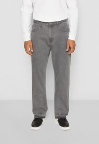 Pantalones de mezclilla gris de corte holgado, con bolsillos laterales y cierre de botón, combinados con zapatillas negras. La tela tiene una ligera textura.