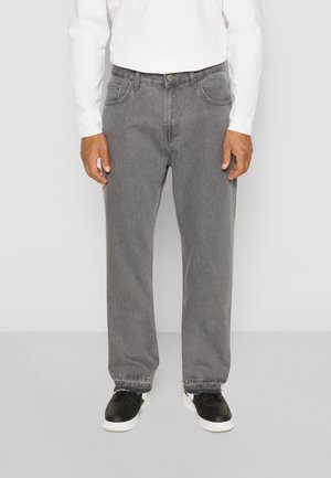 Grijze denim broek met een losse pasvorm, voorzien van zijzakken en een knoopsluiting, gecombineerd met zwarte sneakers. De stof heeft een lichte textuur.