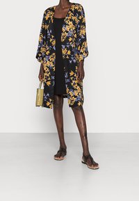 Zwarte kimono-stijl jas met een geel en paars bloemenpatroon, losse pasvorm, halve mouwen, gecombineerd met een zwarte jurk en een doorzichtige tas.