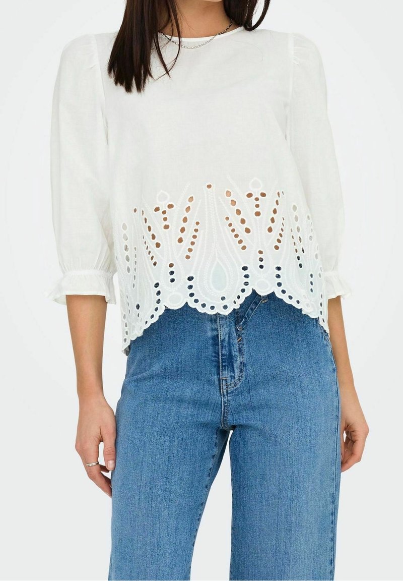 Weiße Bluse mit Dreiviertelärmeln, featuring intricaten Lochstickereien entlang dem Saum und einem lockeren Schnitt. Kombiniert mit blauen Jeans.