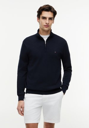 Joven vestido con un suéter azul marino con cremallera hasta el cuello sobre una camisa blanca y pantalones cortos blancos, de pie con una mano en el bolsillo.