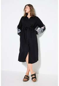 Schwarzes Kleid mit durchgehender Knopfleiste, taillierten Bund und langen Ärmeln, verziert mit kontrastierenden weißen Spitzenmustern. Kombiniert mit schwarzen Sandalen.