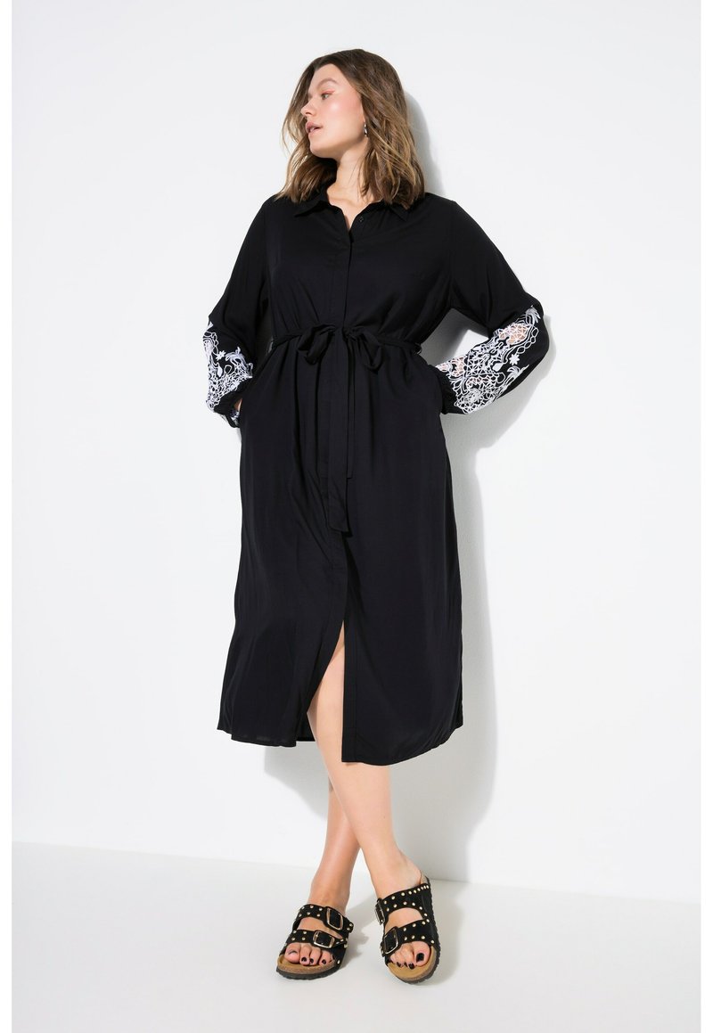 Schwarzes Kleid mit durchgehender Knopfleiste, taillierten Bund und langen Ärmeln, verziert mit kontrastierenden weißen Spitzenmustern. Kombiniert mit schwarzen Sandalen.