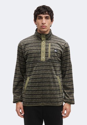 Joven vestido con un jersey verde con estampado, con botones a presión y bolsillos laterales, de pie frente a un fondo claro y liso.