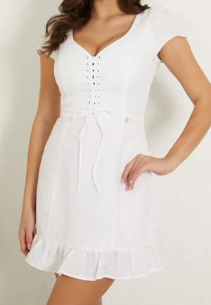 Kobieta ubrana w krótką białą sukienkę z wiązaniem z przodu, krótkimi rękawami i falbaną u dołu, stojąca z jedną ręką na biodrze.