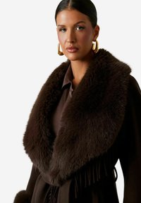Cappotto marrone con colletto in pelliccia morbida, dal design avvolgente e dettagli sfrangiati. Indossato sopra una camicetta marrone, con orecchini dorati e audaci.