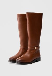 Bottes en cuir marron hauteur genou avec une texture lisse, bout pointu, talon plat et une boucle dorée en accent sur la bride de la cheville.