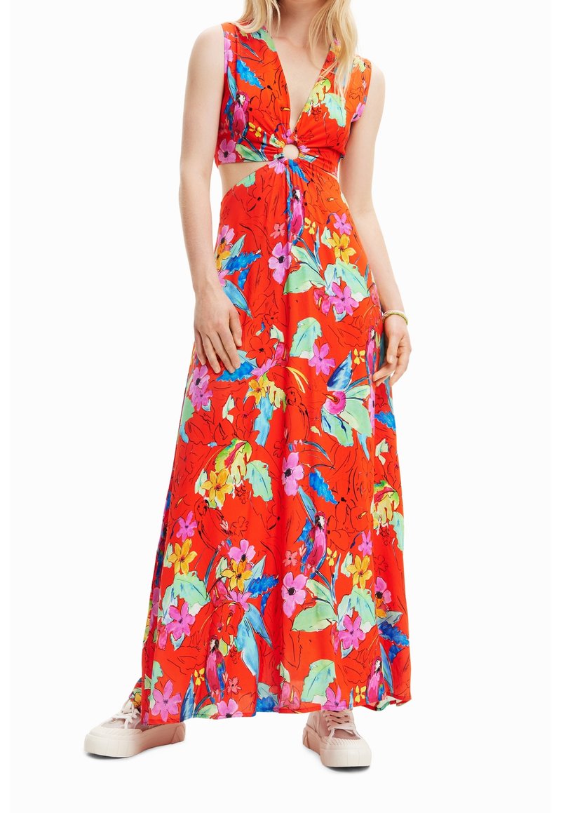 Femme portant une robe maxi rouge sans manches avec un imprimé floral coloré et une découpe sur le devant, assortie à des baskets rose clair.
