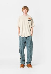 Camiseta de algodón beige con un parche que presenta un perro, combinada con pantalones cargo de denim azul sueltos y calzado marrón con una suela gruesa.