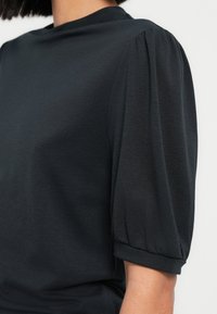 Top grigio scuro con collo alto, maniche corte a sbuffo e una texture liscia e morbida. Il design enfatizza una vestibilità comoda.