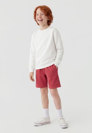 Garçon roux souriant portant un sweat blanc, un short rouge, des chaussettes blanches et des baskets blanches, debout avec une main dans la poche.