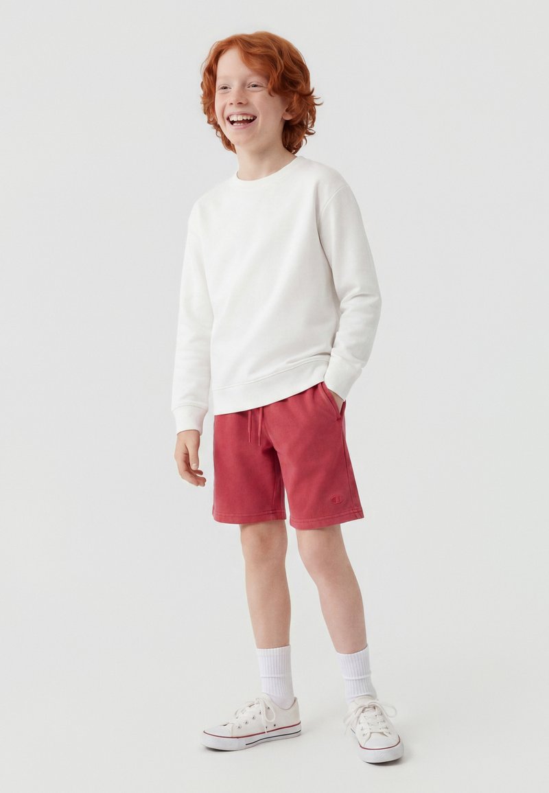 Garçon roux souriant portant un sweat blanc, un short rouge, des chaussettes blanches et des baskets blanches, debout avec une main dans la poche.