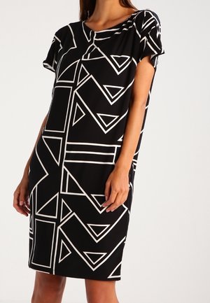 Woman wearing a knee-length black dress with white geometric triangle and square patterns, short sleeves, and a round neckline.