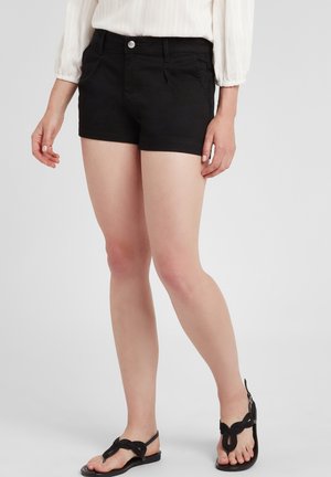Mujer con pantalones cortos negros, blusa blanca de manga larga y sandalias negras, de pie frente a un fondo claro y liso.