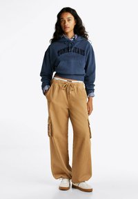 Felpa cropped blu navy con logo "TOMMY JEANS", abbinata a pantaloni cargo beige a gamba larga con tasche laterali e vita regolabile con cordino. Sneakers bianche.