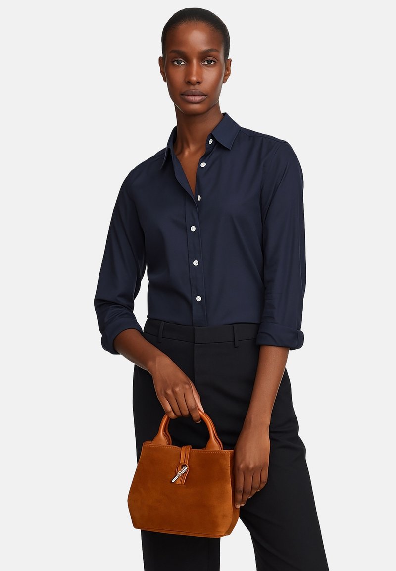 Borsa a tracolla in suede marrone con design strutturato, manici in pelle corti e dettaglio con zip. Modella che indossa una camicia blu navy con bottoni e pantaloni neri.