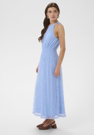 Žena u plavoj maxi haljini bez rukava od čipke, nosi smeđe cipele s niskom blok petom, zlatne naušnice u obliku obruča i zlatne narukvice, stoji bočno.