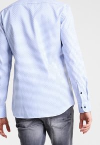 Světle modrá kostkovaná košile s dlouhým rukávem, s manžetami na knoflíky, zakřiveným lemem a kontrastními knoflíky; nošená s šedými džínami z denimu.