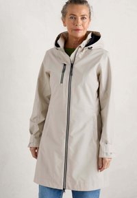 Manteau imperméable beige avec fermeture éclair à l'avant, capuche ajustable et poches latérales. Fabriqué dans un tissu lisse avec une coupe décontractée et des accents aux poignets.