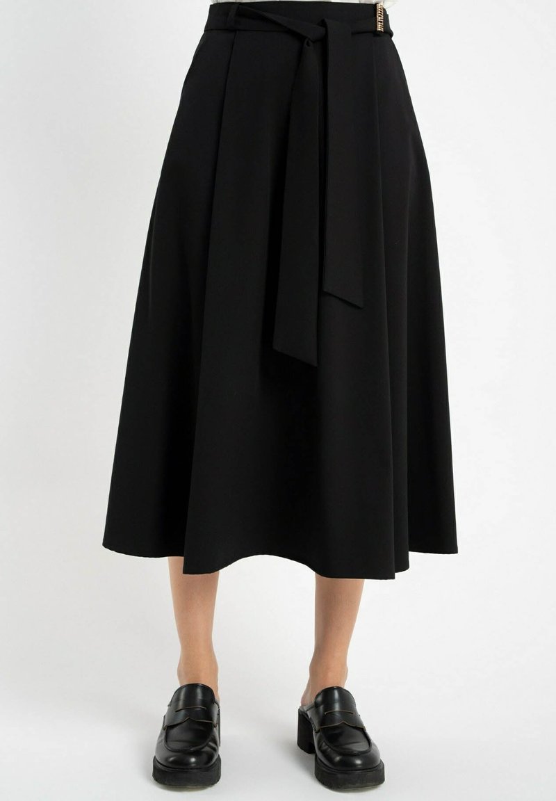 Czarna spódnica do kolan w zwiewnym fasonie z paskiem w talii i bocznymi wiązaniami. Gładki materiał z delikatnym połyskiem. Noszona z czarnymi mokasynami.