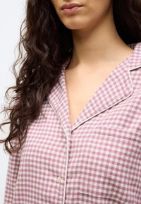 Γυναίκα που φοράει ροζ και λευκό καρό πιτζαμάκι με λευκές ραφές και τσέπη στο μπροστινό μέρος, με μαλλιά μέχρι τους ώμους και σγουρά.