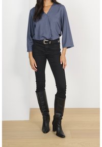 Femme portant un chemisier bleu texturé, un pantalon skinny noir avec une ceinture de style western, et des bottes en cuir noires à talons montant jusqu'au genou.