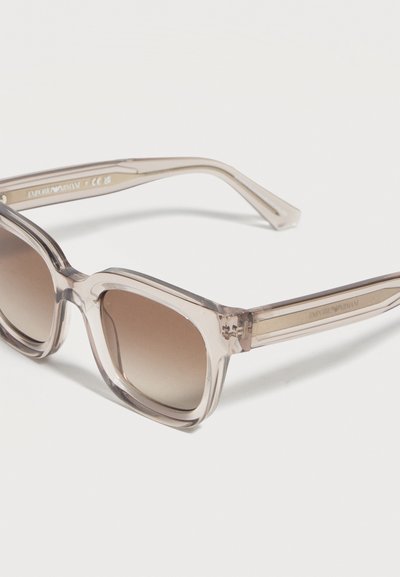 Διαφανή μπεζ υπερμεγέθη τετράγωνα γυαλιά ηλίου με καφέ φακούς με διαβάθμιση και χρωματιστούς χοντρούς βραχίονες σε λευκό φόντο.