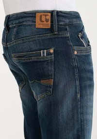 Jeans de mezclilla azul oscuro con un parche de cuero, múltiples bolsillos y costuras en contraste. La tela tiene una textura suave con un desgaste mínimo.