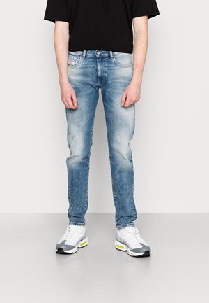 Person, der verblasste blaue Jeans und weiße-graue Turnschuhe trägt, steht vor einem schlichten hellen Hintergrund.
