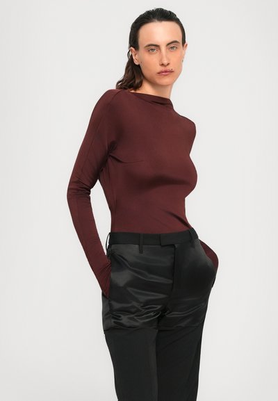 Woman posing with hands in pockets, wearing a fitted long-sleeve burgundy top and black pants with shiny fabric on the upper thighs.