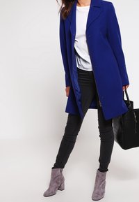 Dlouhý modrý kabát s klopovým límcem a dvěma předními knoflíky, v kombinaci s černými skinny džíny a šedými kotníkovými botami. Souč�ástí je černá taška.