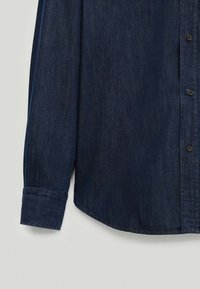 Donkerblauw denim overhemd met lange mouwen, knoopmanchet en zichtbare knoopsluiting op een lichtgrijze achtergrond.