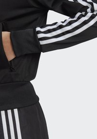 Chaqueta negra con detalle de tres rayas blancas en las mangas. Cuenta con un bolsillo con cremallera y puños acanalados, fabricada en un tejido suave y liso.