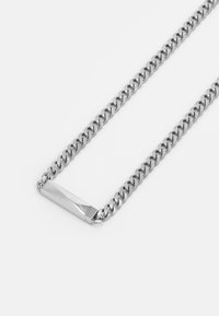 Sølvfarget metallkjede med sammenkoblede lenker, med en elegant rektangulær anheng som viser et merkevarelogo. Glatt tekstur.