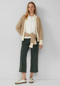 Strickjacke im braunen und cremefarbenen Hahnentrittmuster, getragen über einem weißen Hemd, mit grünen, knöchelfreien Hosen und beigen Sneaker-Akzenten.