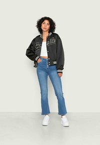 Casaco bomber preto de satin com detalhes brancos, top cropped branco, jeans azul de cintura alta com detalhe de botão e ténis brancos.