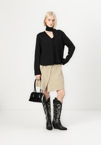 Top nero a coste con bottoni e scollatura a V, gonna avvolgente beige, borsa nera e stivali al ginocchio neri con ricami floreali bianchi.