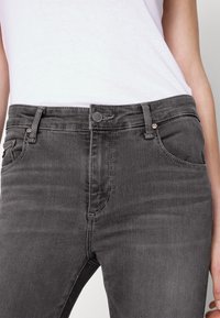 Jeans gris oscuro con un diseño ajustado, estilo clásico de cinco bolsillos, y detalles de costura visibles; cuenta con un cierre de botón y cremallera.