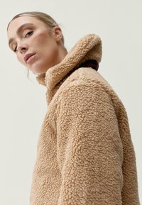 Béžová fleecová bunda s vysokým límcem a texturovaným povrchem, s předním zipem a bočními kapsami, umístěná na neutrálním pozadí.