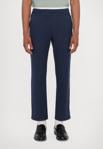 Pantalon bleu marine à coupe droite, avec une taille élastique et une fermeture à bouton. Fabriqué en tissu texturé, associé à des mocassins noirs et des chaussettes blanches.